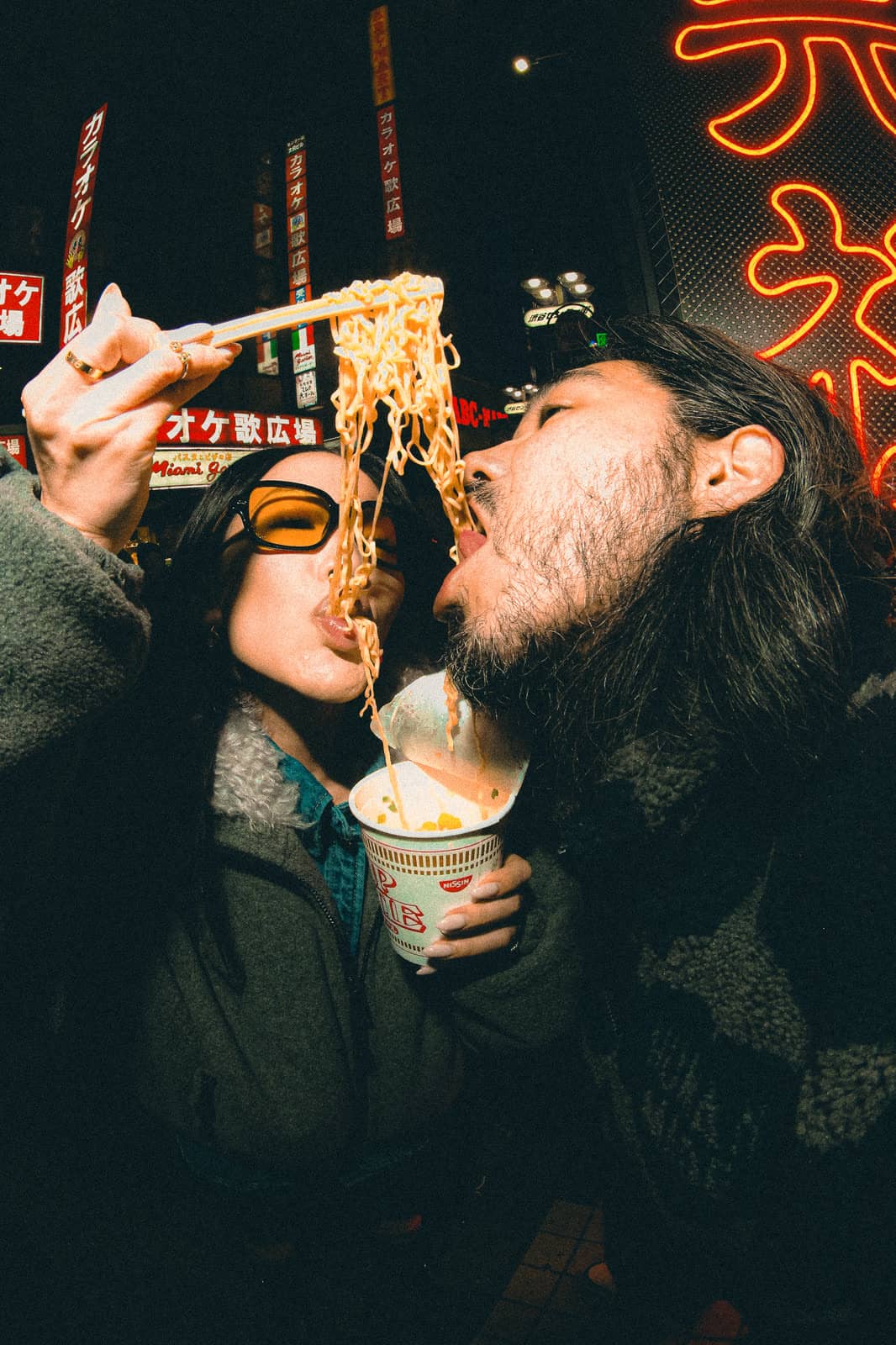 Couple sharing a cup of noodles on a neon street at night